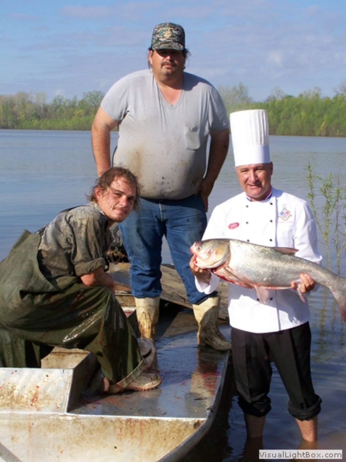 Chef Philippe Parola holding a large carp alongside two fishermen beside a river
